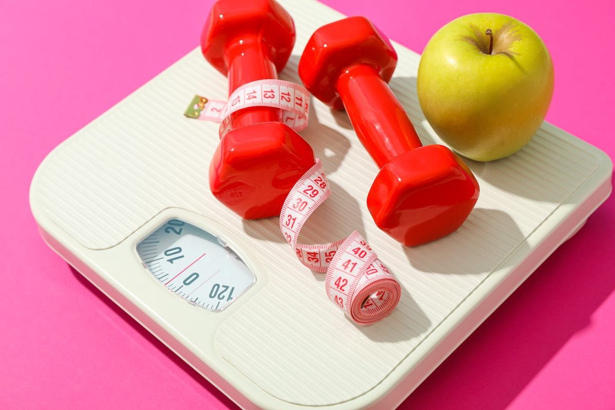 Weigh scales, apple, dumbbells and measuring tape on pink background