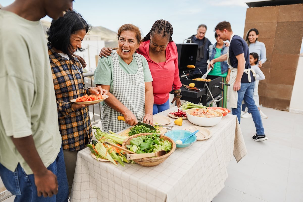 Multi generational people doing barbecue at home’s rooftop – Multiracial friends having fun eating and cooking together during weekend day – Summer and food concept – Main focus on latin woman face